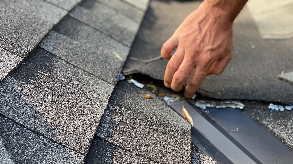 Roofer on a ladder replacing blown-off asphalt shingles along a gutter line